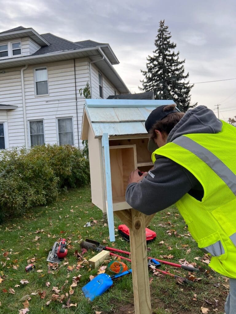 A WMCI student installs a little library they built in their carpentry class. He's placing it in front of a house in the Roosevelt Park neighborhood of Grand Rapids, MI.