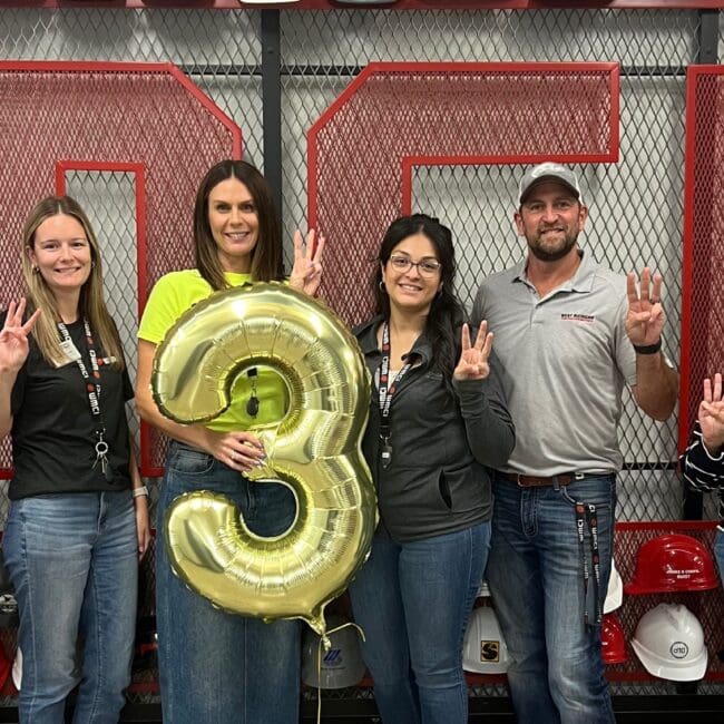 The full WMCI team stands in front of the hard hat donor wall with a '3' balloon marking the 3rd year in business.