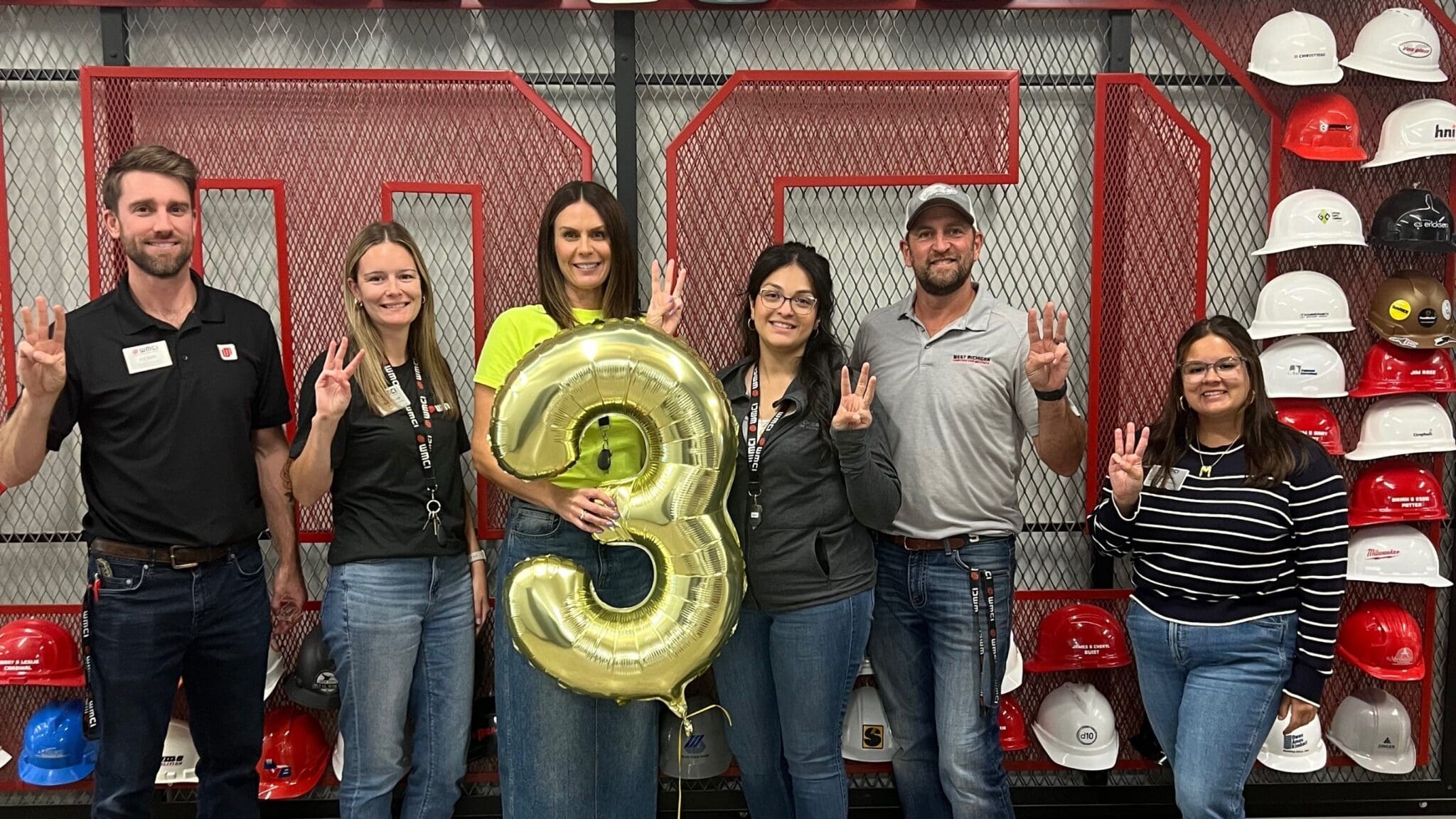 The full WMCI team stands in front of the hard hat donor wall with a '3' balloon marking the 3rd year in business.