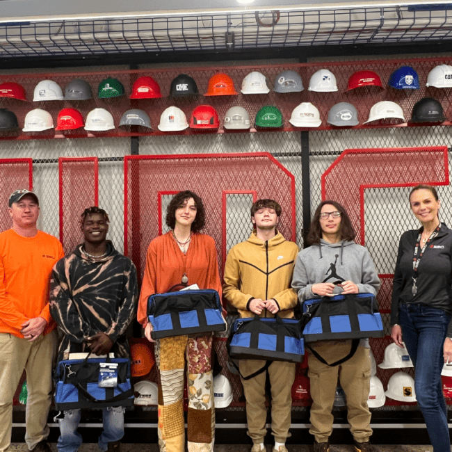 A group of 5 WMCI work based learning students celebrate graduation in front of the West Michigan Construction Institute hard hat wall.